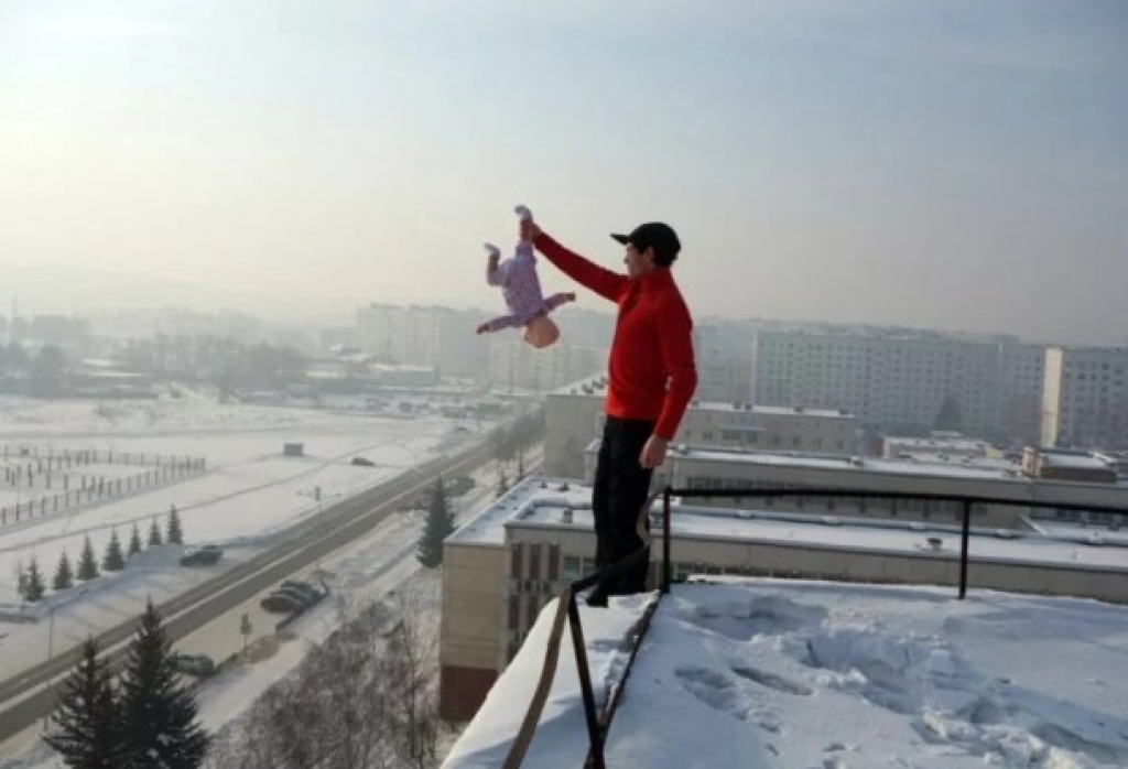 Russo posa na borda do telhado segurando filha bebê (ou boneca?) de cabeça para baixo - Foto: Siberian Times